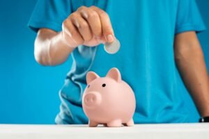A close-up of an adult's hand dropping a coin into a piggy bank, symbolizing savings and investment.