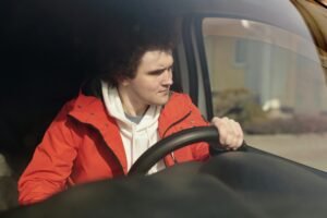 A young man in a red jacket steering a car with focus, enjoying a peaceful drive.